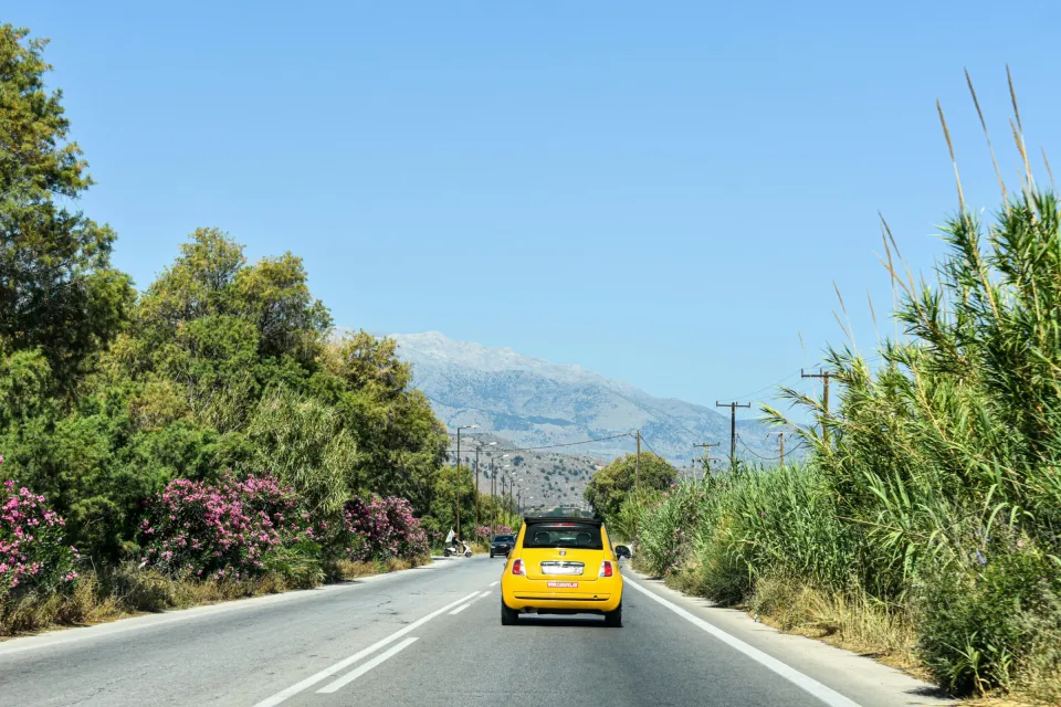 Yellow car drives down a scenic road in Greece, surrounded by lush greenery and distant mountains.